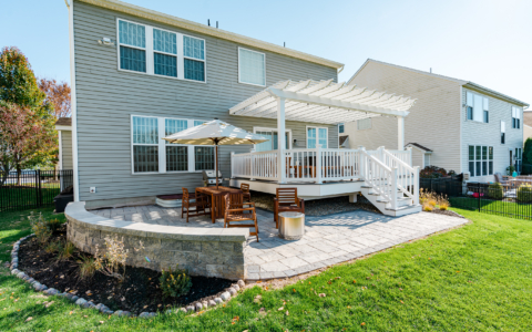 image of a deck and patio in downingtown pa built by integrous fences and decks featuring a white vinyl pergola timbertech decking white vinyl railing paver patio and sitting wall around the yard is a black aluminum fence